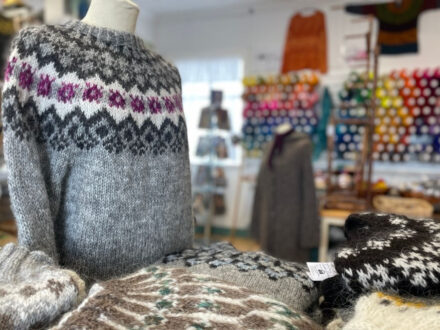 A display of traditional Icelandic lopapeysa sweaters, with one on a mannequin featuring a gray and white pattern with a vibrant purple design, and several others folded on a table in the foreground.