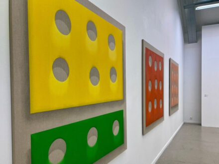 Contemporary art exhibit with brightly colored rectangular artworks, including a prominent yellow piece resembling cheese with holes, and a smaller green piece, against a plain gallery wall."