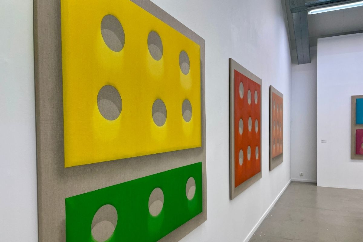 Contemporary art exhibit with brightly colored rectangular artworks, including a prominent yellow piece resembling cheese with holes, and a smaller green piece, against a plain gallery wall.