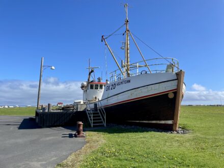 The fishing vessel GK 20 Holsteinn on display outdoors at the Heritage Museum of Garðskagi, with a gangway leading to its deck and green fields in the background under a blue sky.