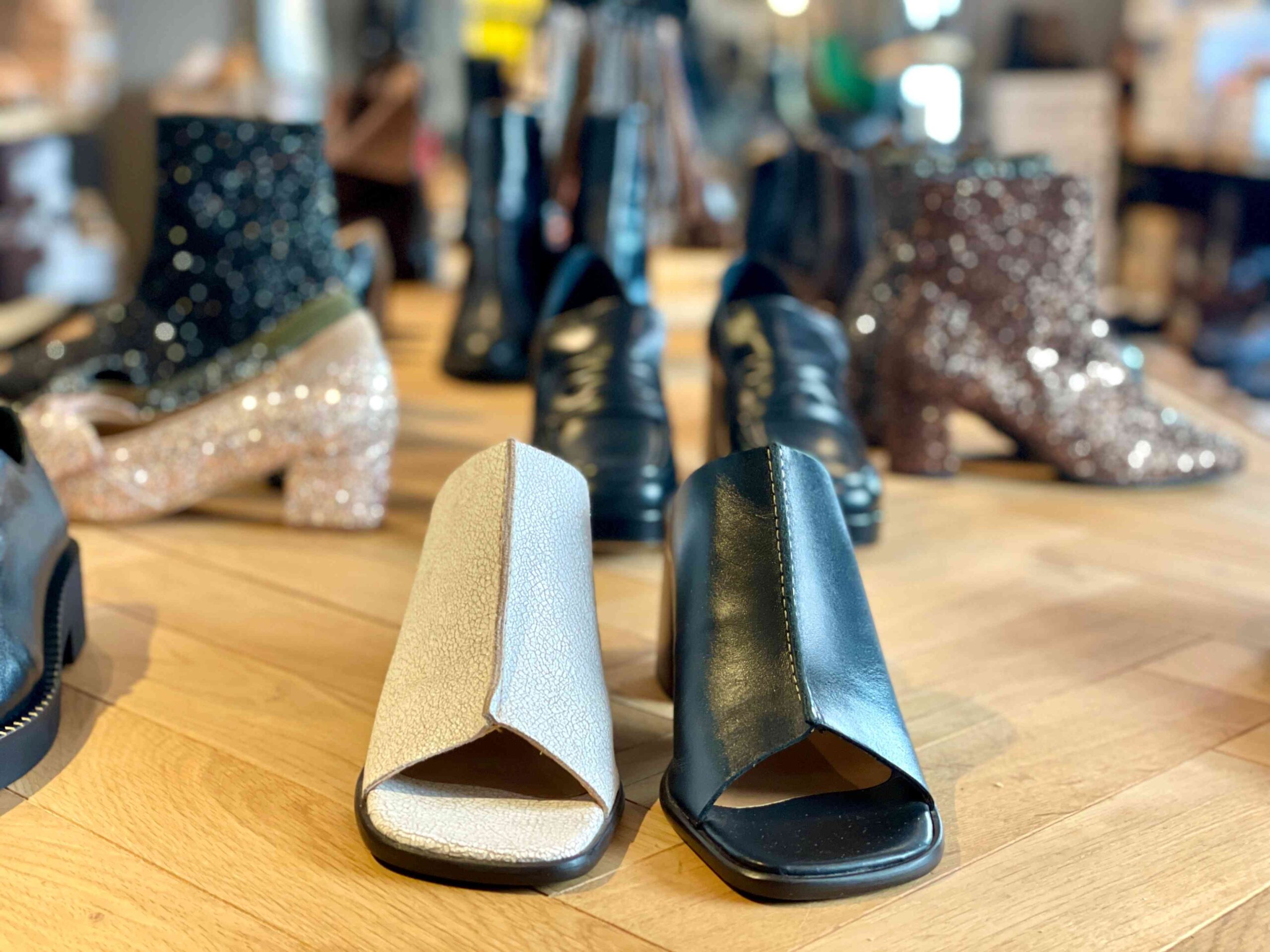 A pair of open-toe, block-heeled shoes in light grey and black, with other classy shoes in the background, at Fló shoes.