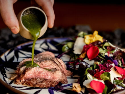 A beautifully presented dish at Kol Restaurant in Reykjavik, featuring a lamb steak with a vibrant green sauce being poured over it, accompanied by a colorful salad.