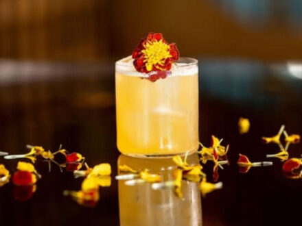 Elegant floral cocktail from Monkeys Restaurant in Reykjavik, garnished with edible red and yellow flowers on a black reflective surface.