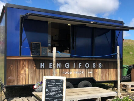 The "Hengifoss Food Truck," a blue and wood-paneled trailer with an open service window and a "MENU" chalkboard in front, parked on a grassy area under a partly cloudy sky.