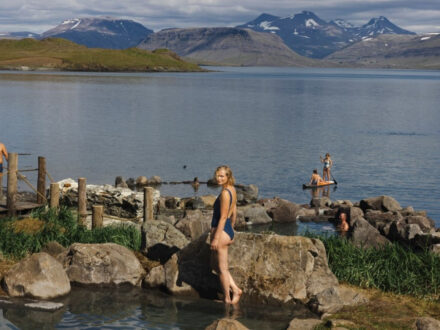People relaxing in and around outdoor geothermal pools next to a vast, calm lake, with snow-capped mountains under a partly cloudy sky in the background. One woman stands on a rock in the foreground, and others are in the water and on a paddleboard.