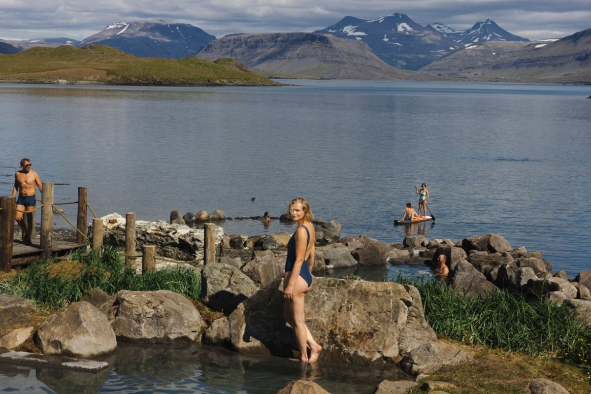 People relaxing in and around outdoor geothermal pools next to a vast, calm lake, with snow-capped mountains under a partly cloudy sky in the background. One woman stands on a rock in the foreground, and others are in the water and on a paddleboard.