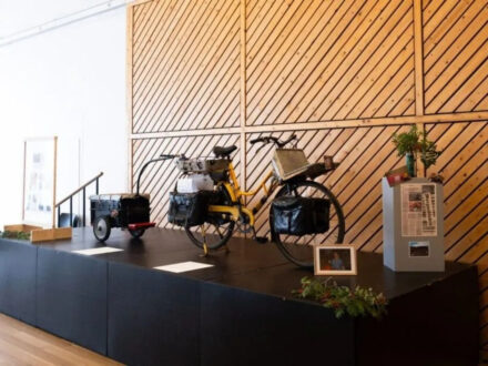 A museum display at the Technical Museum of East Iceland featuring a historic bicycle with attached luggage and a small trailer, surrounded by other exhibit elements and a modern wooden wall.