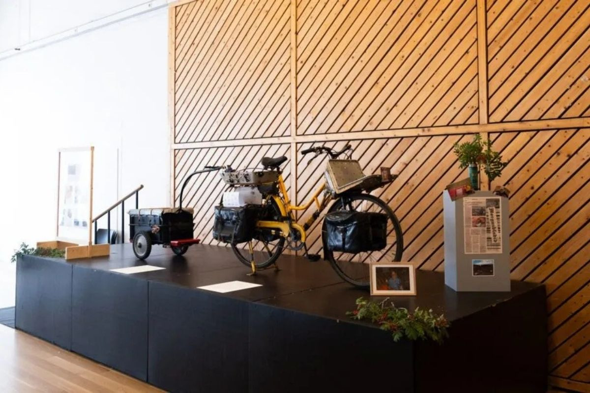 A museum display at the Technical Museum of East Iceland featuring a historic bicycle with attached luggage and a small trailer, surrounded by other exhibit elements and a modern wooden wall.
