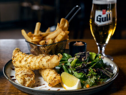 A plate of crispy fish and chips with a side salad and a lemon wedge, accompanied by a basket of fries and a tall glass of Gull beer, likely from Vegamót Bíldudal in Iceland.