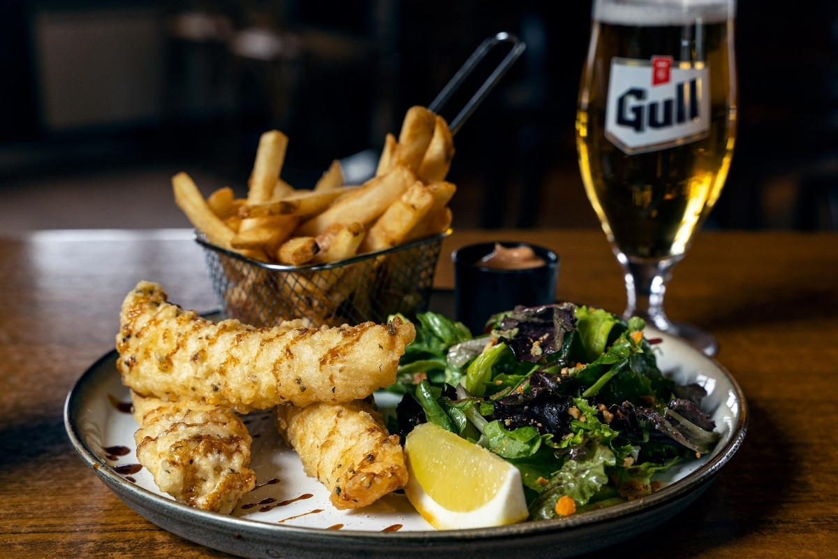 A plate of crispy fish and chips with a side salad and a lemon wedge, accompanied by a basket of fries and a tall glass of Gull beer, likely from Vegamót Bíldudal in Iceland.