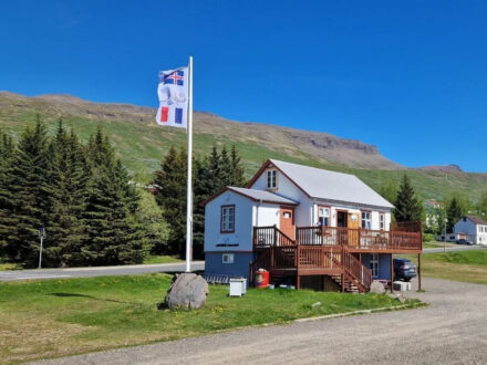 Café Sumarlína, a white building with a distinctive brown deck, stands under a clear blue sky in rural Iceland, marked by a prominent French flag.