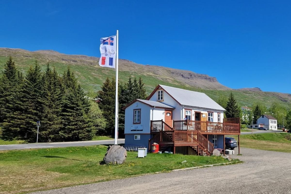 Café Sumarlína, a white building with a distinctive brown deck, stands under a clear blue sky in rural Iceland, marked by a prominent French flag.
