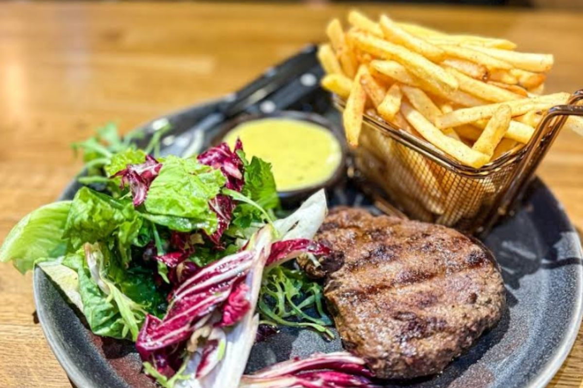 A grilled burger patty served with a side of crispy french fries in a metal basket and a fresh green salad with a small bowl of sauce, at Public Deli Restaurant.