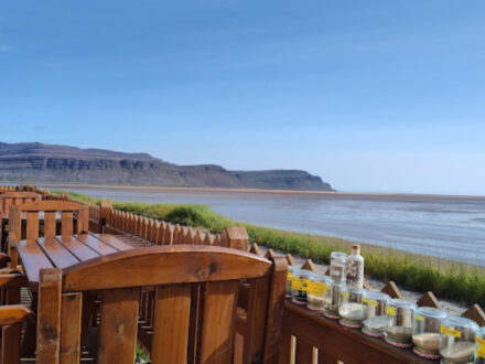 An outdoor dining area at The French Café in Rauðasandur, Iceland, with wooden tables and chairs facing a wide bay with a low tide and distant mountains under a clear blue sky. Jars with various contents are displayed on the railing.