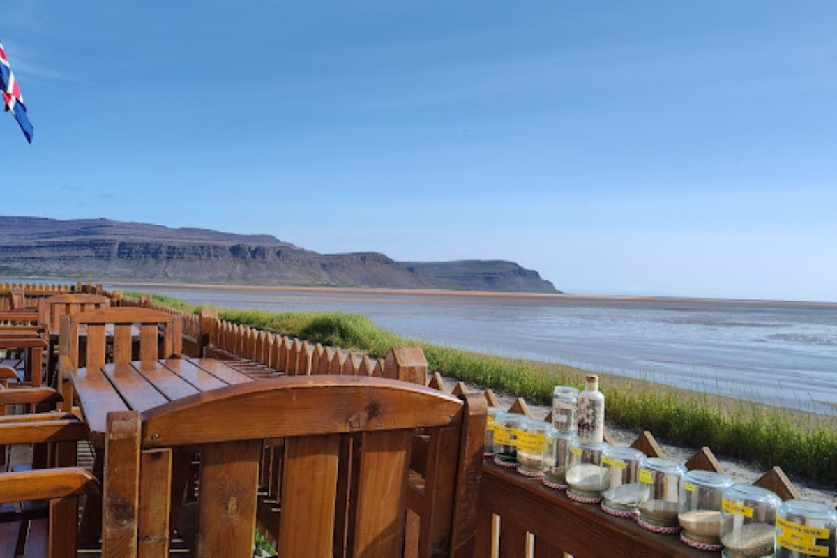 An outdoor dining area at The French Café in Rauðasandur, Iceland, with wooden tables and chairs facing a wide bay with a low tide and distant mountains under a clear blue sky. Jars with various contents are displayed on the railing.