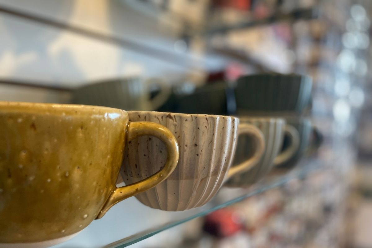 A close-up, eye-level shot of handcrafted ceramic mugs in various earthy tones, including speckled gold and a textured cream color, displayed on a glass shelf.