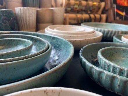 A close-up, low-angle shot of a collection of handcrafted, turquoise-colored ceramic bowls with a textured, speckled glaze, nested on a dark surface in Gallery Flói, Iceland.