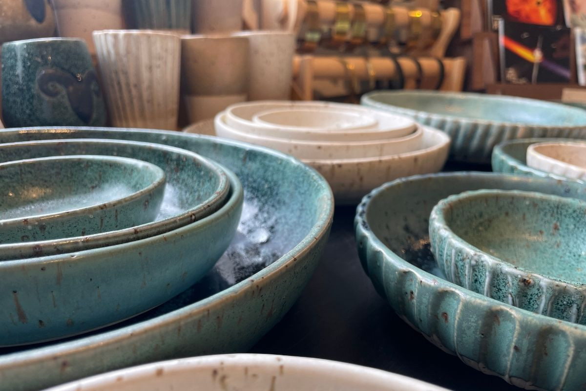 A close-up, low-angle shot of a collection of handcrafted, turquoise-colored ceramic bowls with a textured, speckled glaze, nested on a dark surface in Gallery Flói, Iceland.