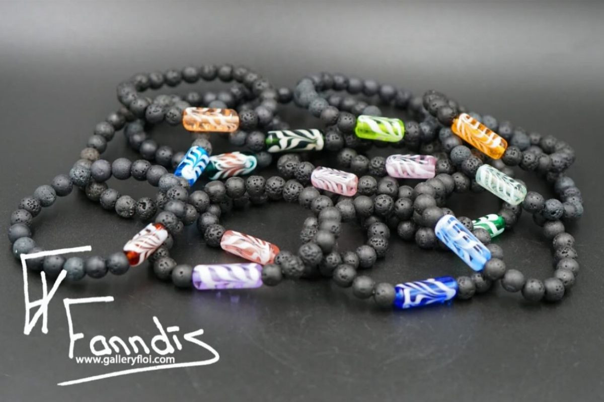 Multiple black beaded bracelets, possibly made of lava beads, each featuring a unique rectangular glass bead in various colors and patterns, arranged on a dark surface, showcasing Icelandic artisan jewelry from Gallery Flói by Fanndís Huld Valdimarsdóttir.