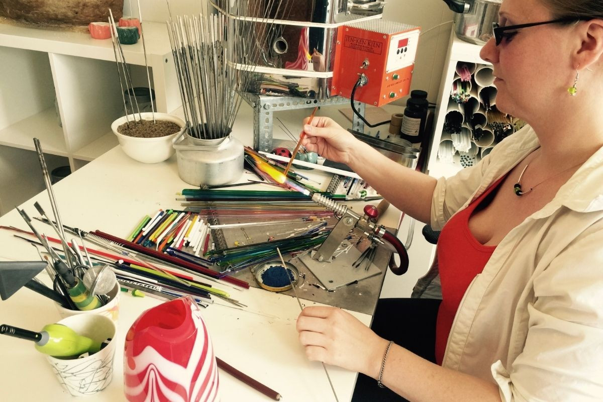 Icelandic artisan Fanndís Huld Valdimarsdóttir at her workbench, wearing glasses and a red top under a white shirt, holding a glass rod and working on a piece of art with various tools and colorful glass rods spread across the table, showcasing her craft at Gallery Flói.