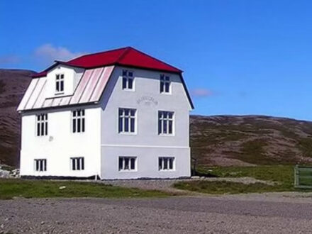 The historic Gamli bærinn Café in Iceland, a distinctive two-story white building with a vibrant red roof, set against a backdrop of rolling brown hills under a clear blue sky.