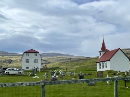 The white church with a red steeple and a nearby traditional Icelandic farmhouse at Gamli bærinn - Brjánslæk, surrounded by a green cemetery and rolling hills under a cloudy sky.