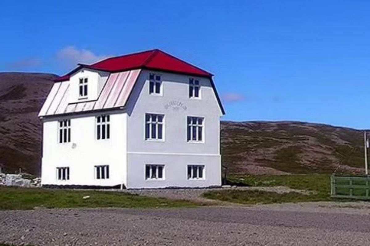 The historic Gamli bærinn Café in Iceland, a distinctive two-story white building with a vibrant red roof, set against a backdrop of rolling brown hills under a clear blue sky.