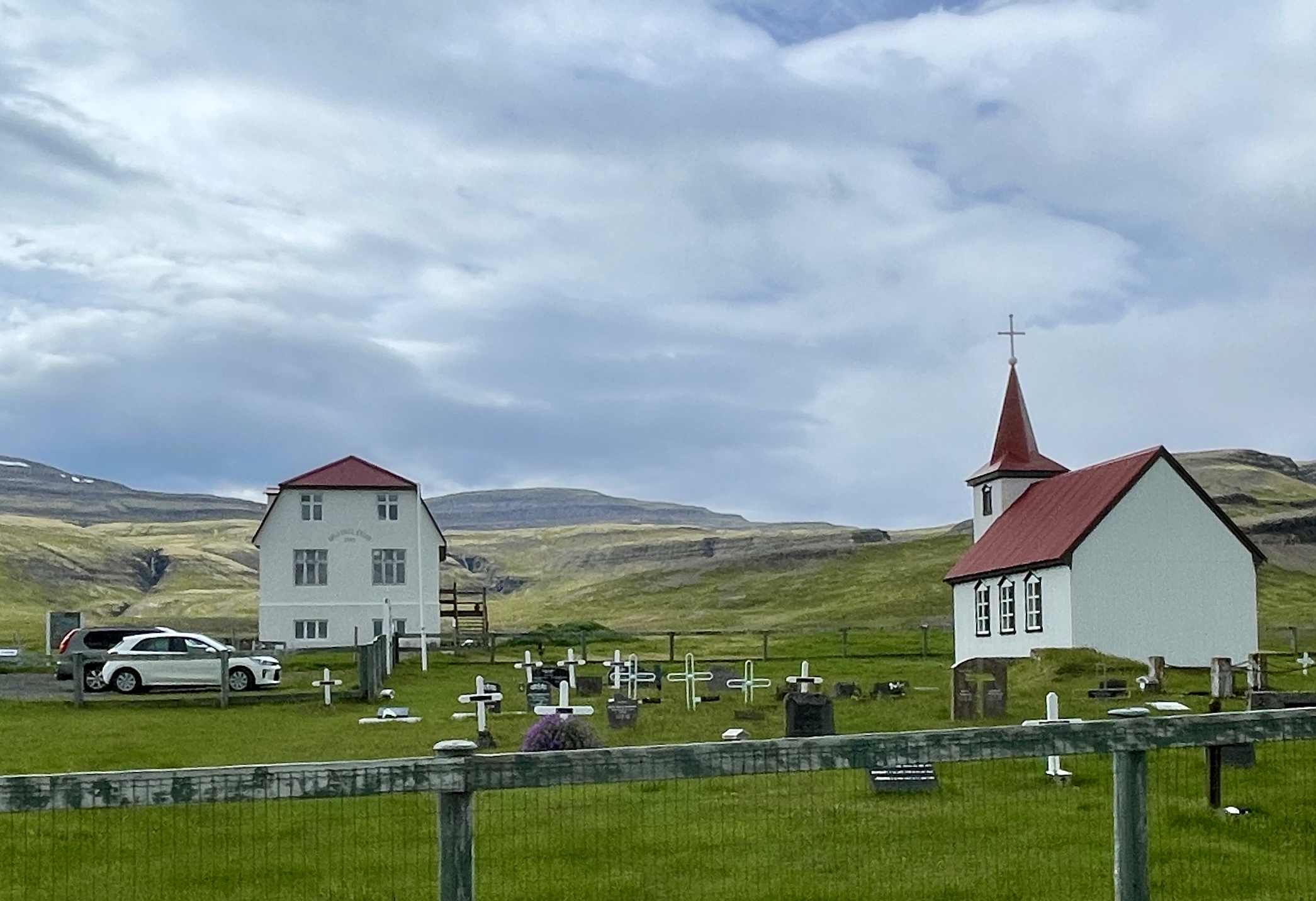 The white church with a red steeple and a nearby traditional Icelandic farmhouse at Gamli bærinn - Brjánslæk, surrounded by a green cemetery and rolling hills under a cloudy sky.