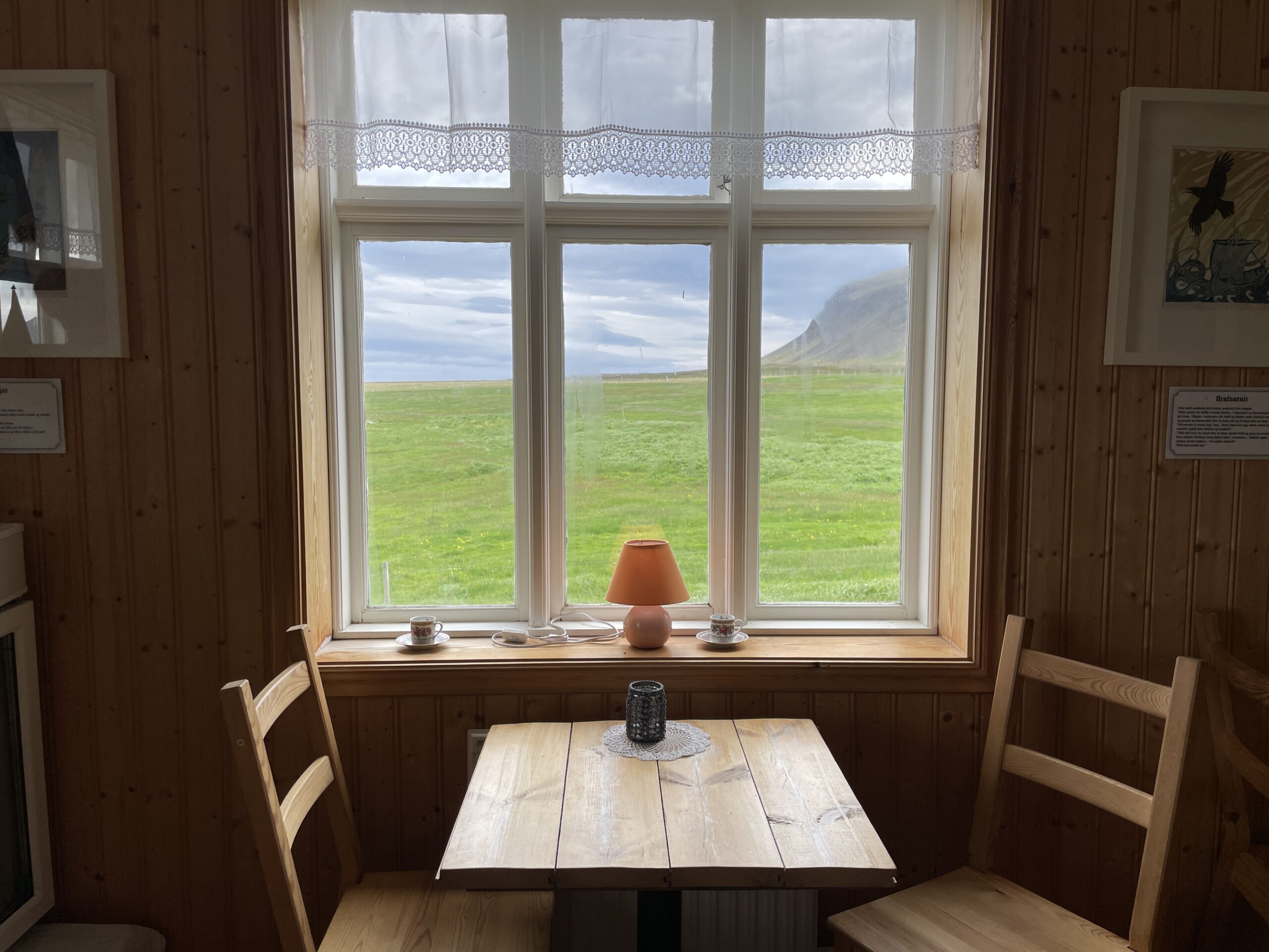 A cozy wooden interior of The Old Farmhouse Cafe (Gamli bærinn) with a table and chairs facing a window overlooking a vast green field and distant mountains in Iceland.