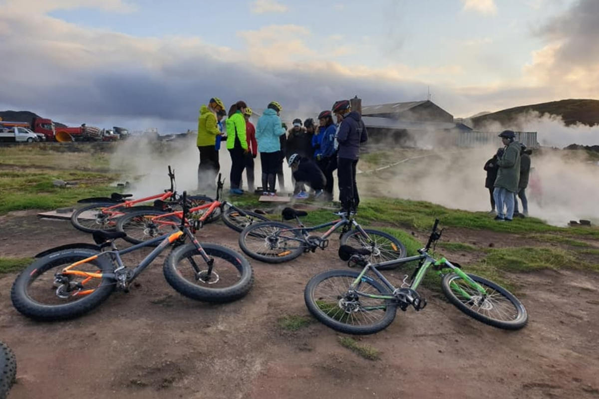 A group of mountain bikers and their bikes gathered in a geothermal area with steam rising from the ground, indicating a bike tour experience near Lake Myvatn in Iceland.