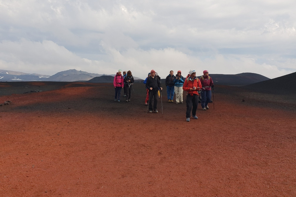 A group of hikers in colorful outdoor gear walking across a vast, reddish-brown volcanic landscape under a cloudy sky, showcasing a hiking tour in the Lake Myvatn area of Iceland.