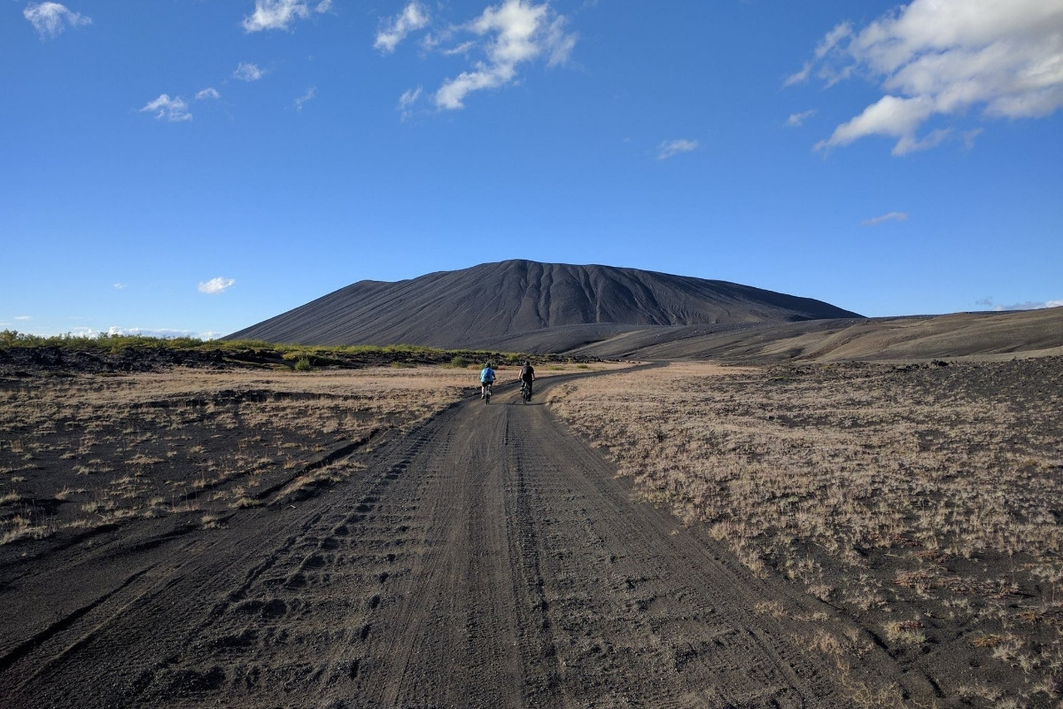 Two cyclists on a dirt road leading towards the large, dark Hverfjall volcano under a blue sky, illustrating a fat bike tour in the Lake Myvatn area of Iceland.