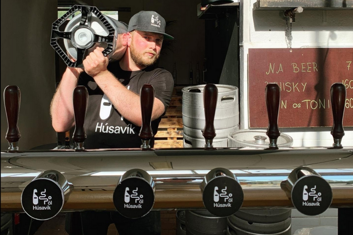 A man at a Húsavík brewery stand with beer taps and a menu.
