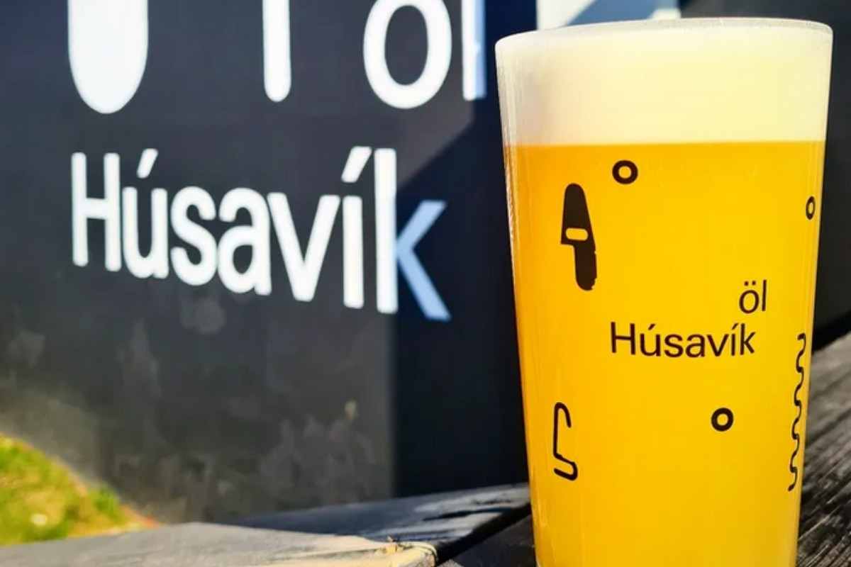A glass of golden beer with foam, displaying the Húsavík Öl logo, next to a black sign.