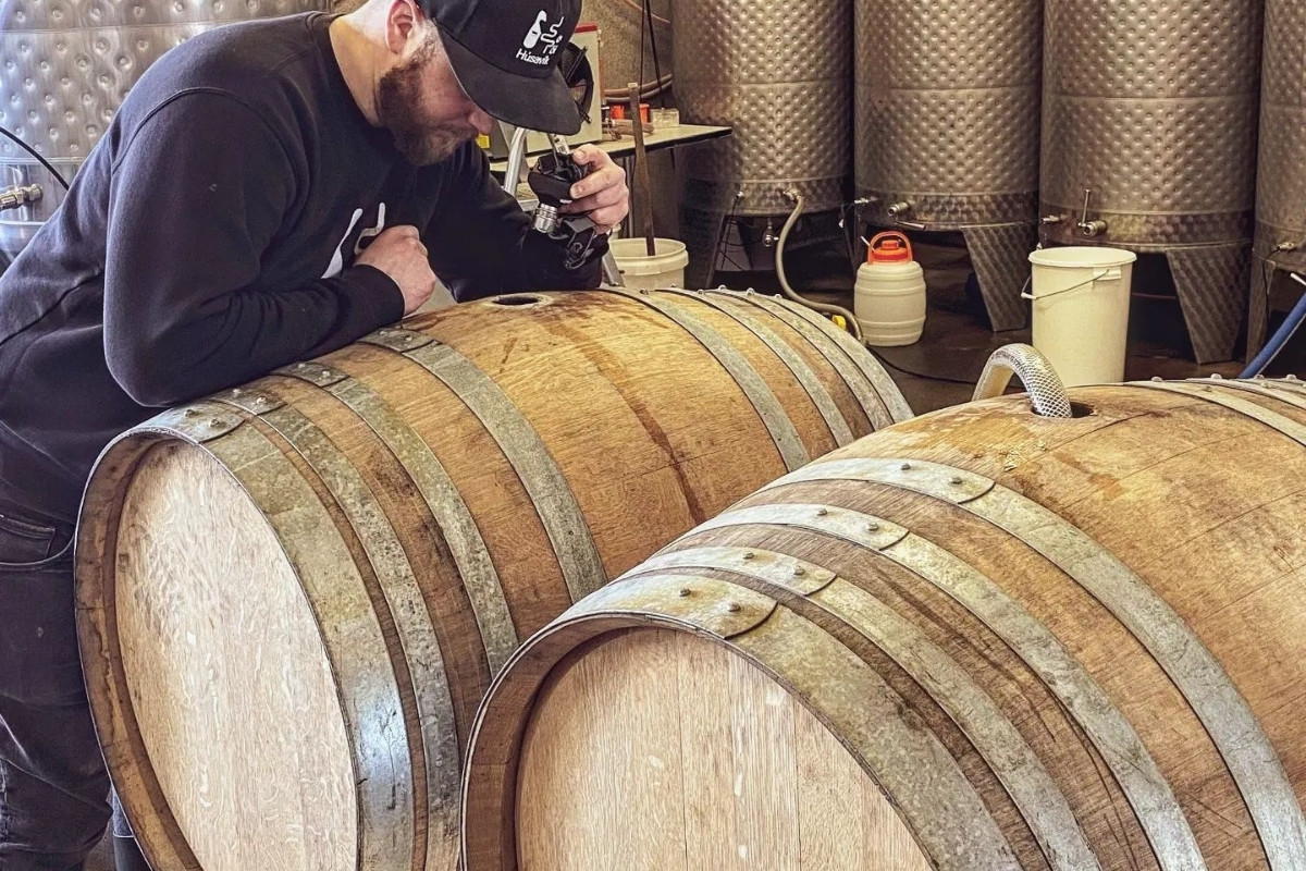 A man inspecting a large wooden barrel in a brewery with metal tanks in the background.