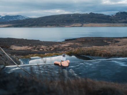 Hvammsvík Hot Springs in Iceland, showing a person soaking in an outdoor hot pool with a stunning view of a fjord and rugged terrain.