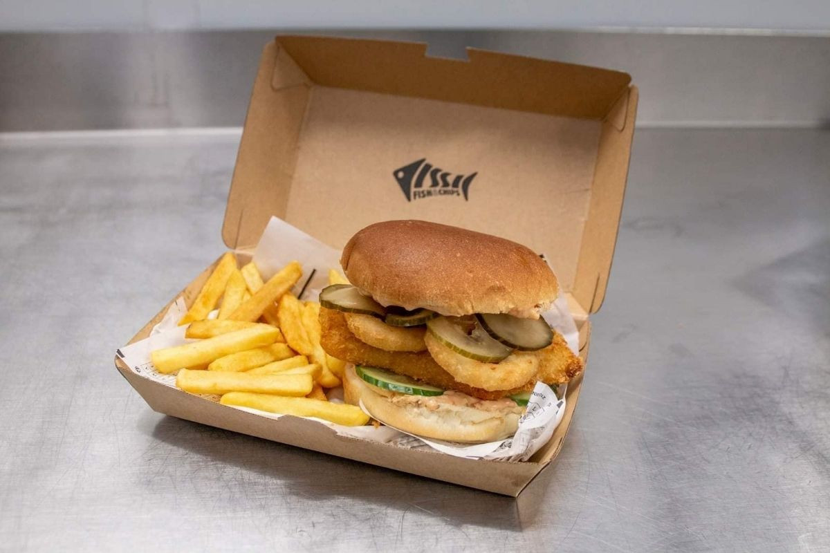 A fish burger with onion rings and pickles, served with fries in an open cardboard box from Issi Fish & Chips.