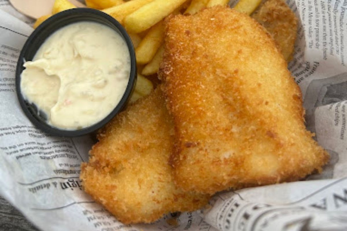 Two pieces of fried fish and french fries, served with a side of white dipping sauce in a black cup, all on paper resembling newspaper.