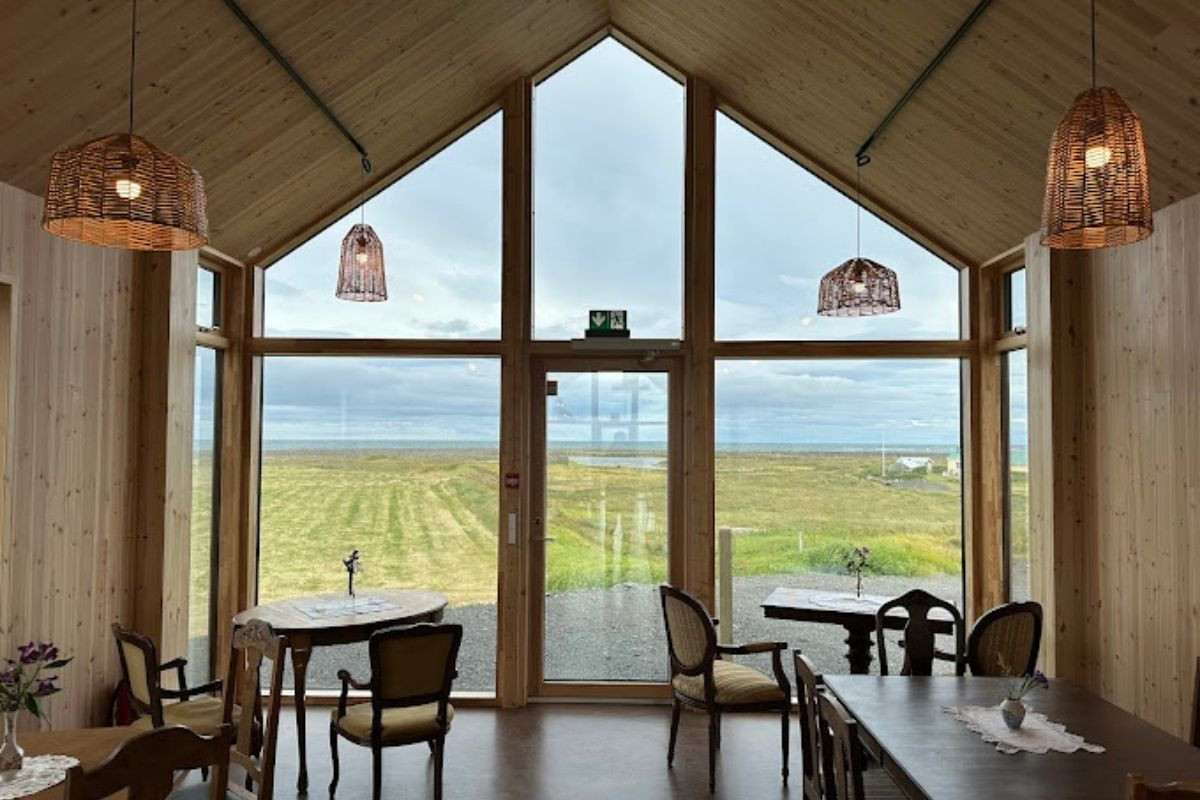 The interior of a cafe with high, wooden-paneled ceilings and large, A-frame windows offering a panoramic view of a vast green landscape under a cloudy sky. Several wooden tables with chairs are arranged throughout the room, and woven pendant lights hang from the ceiling.