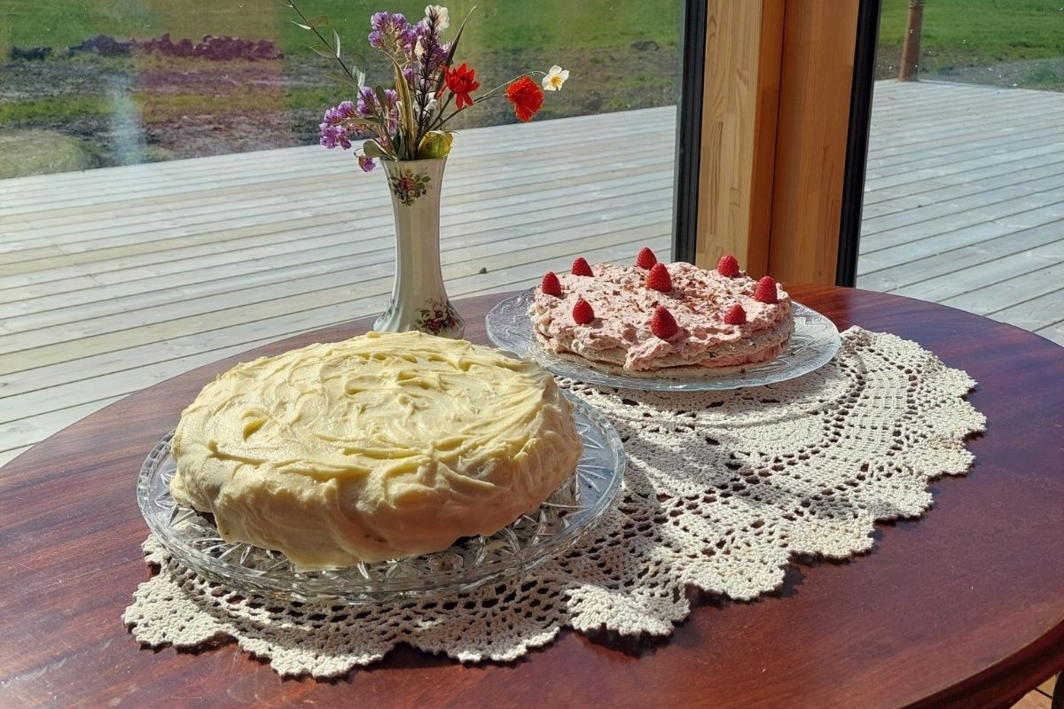 Two cakes, one yellow and one pink with raspberries, on a table with a doily.