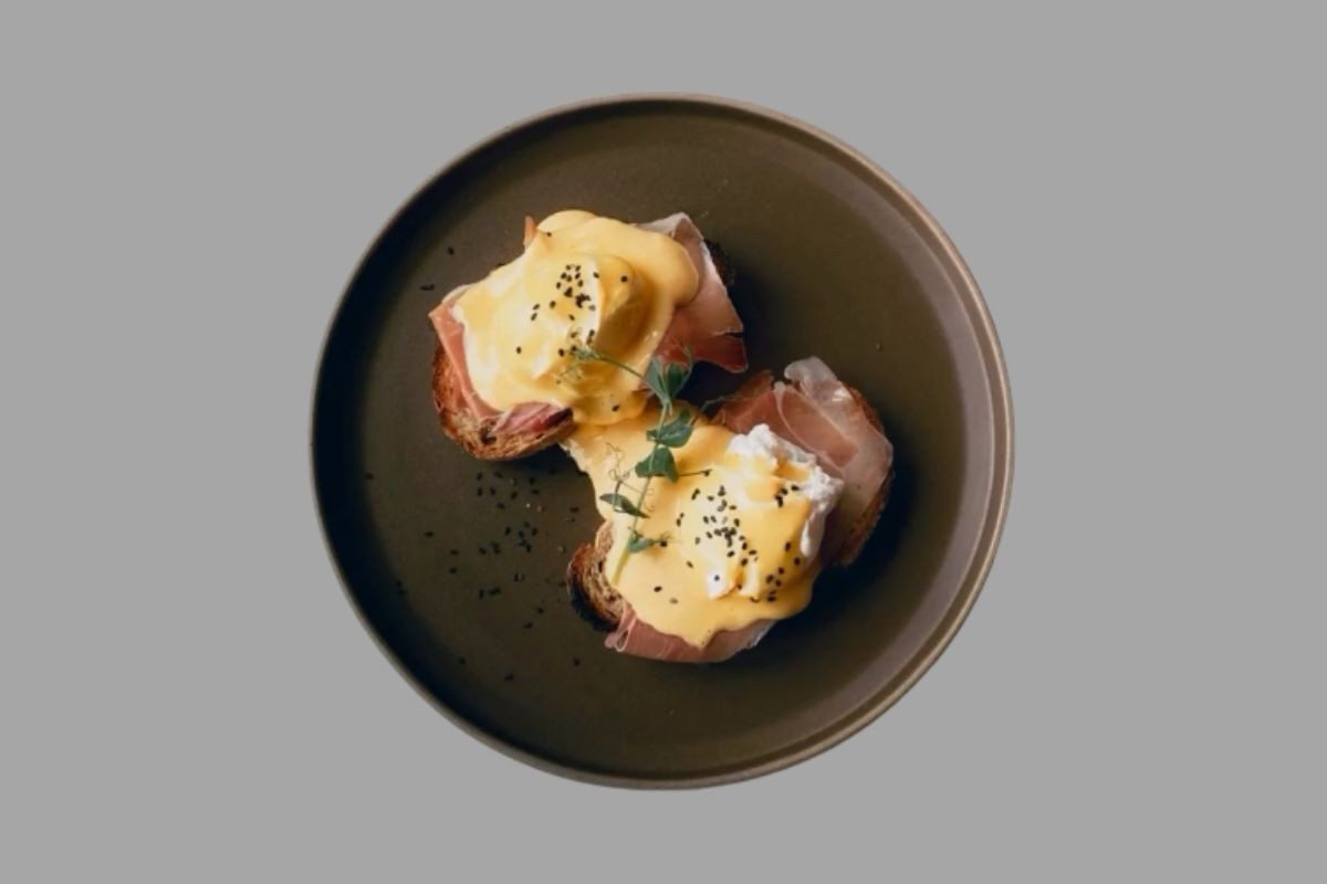 Overhead view of two eggs Benedict with ham on toast, topped with hollandaise sauce and black seeds, on a brown plate.