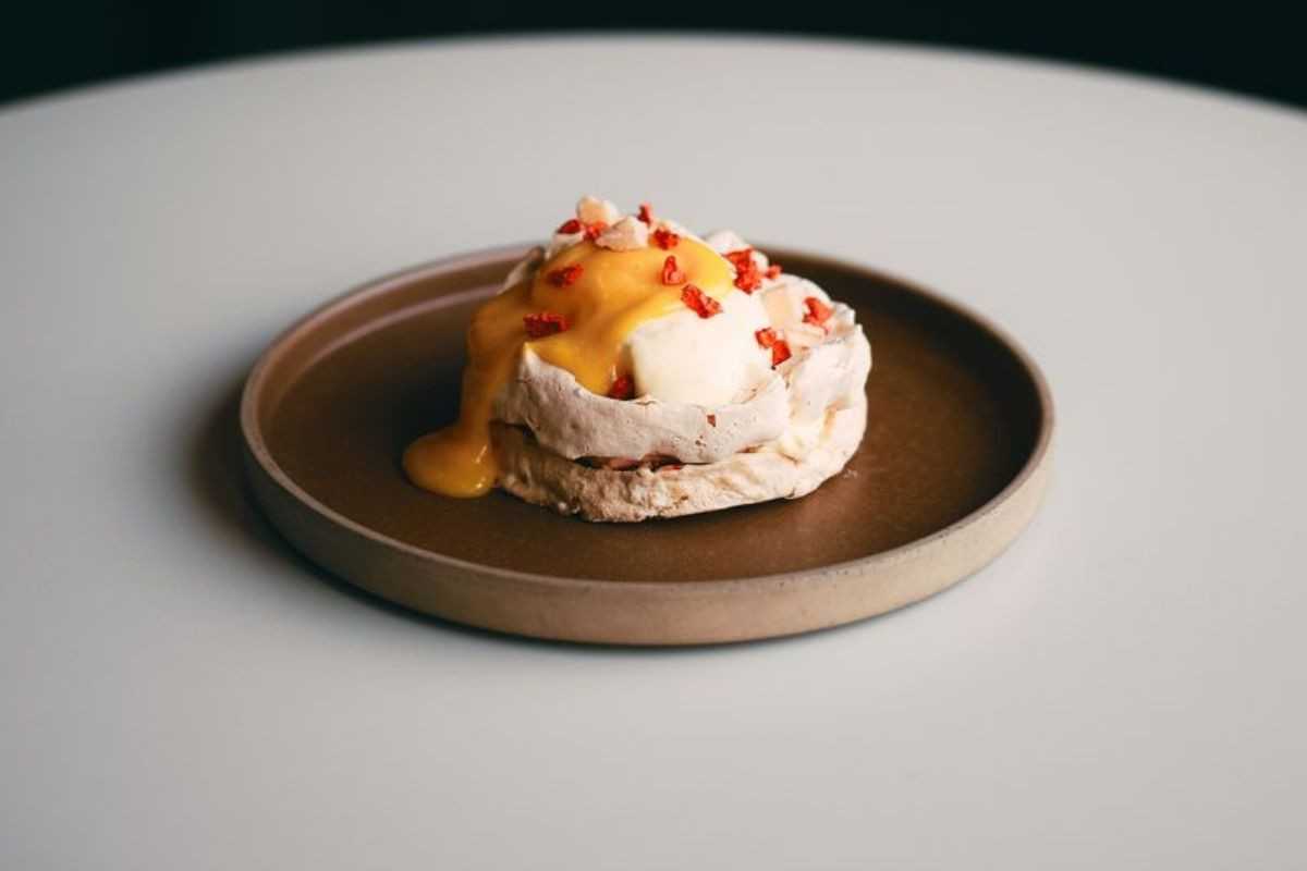 A dessert on a brown plate, consisting of a meringue base, white cream, yellow sauce, and red garnishes.
