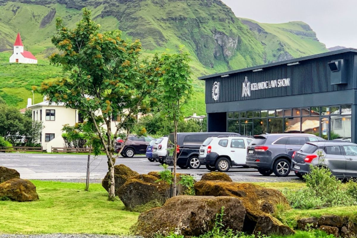 Exterior of the Icelandic Lava Show building with cars in the parking lot and a church on a hill in the background.