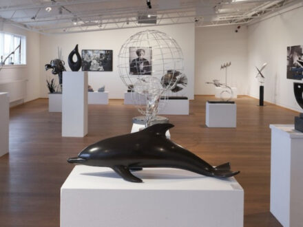 The spacious interior of the Reykjanes Art Museum (Listasafn Reykjanesbæjar), showcasing various contemporary sculptures and artworks on white pedestals, with a large dark dolphin sculpture in the foreground and a spherical wire globe in the middle ground.