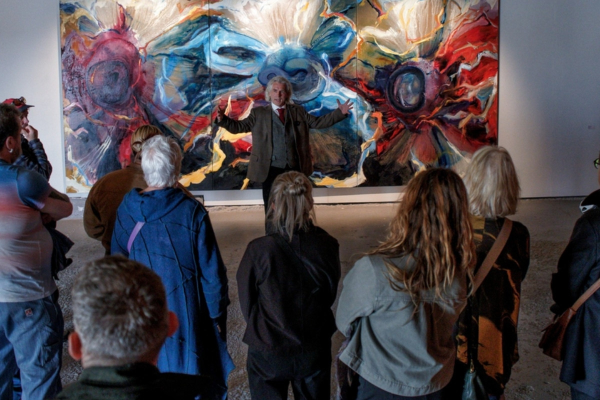 A group of people listening to a speaker in front of a large, colorful abstract painting in an art gallery.