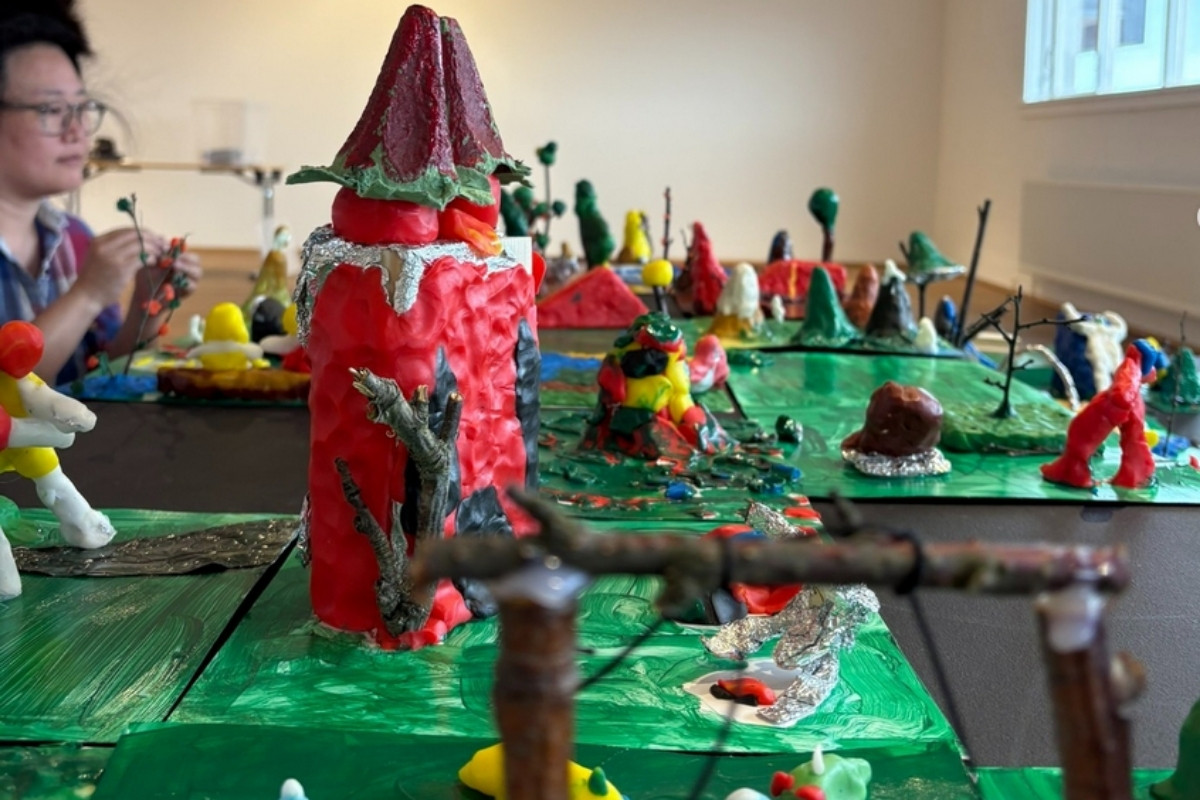 A table covered with various colorful, whimsical clay or mixed-media sculptures depicting a fantastical landscape, with a person in the background.