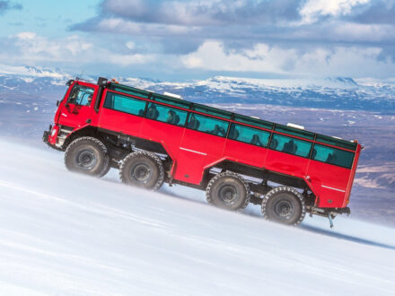 The impressive red Sleipnir monster truck ascending a snowy slope with mountains and a cloudy sky in the background during a glacier tour in Iceland.