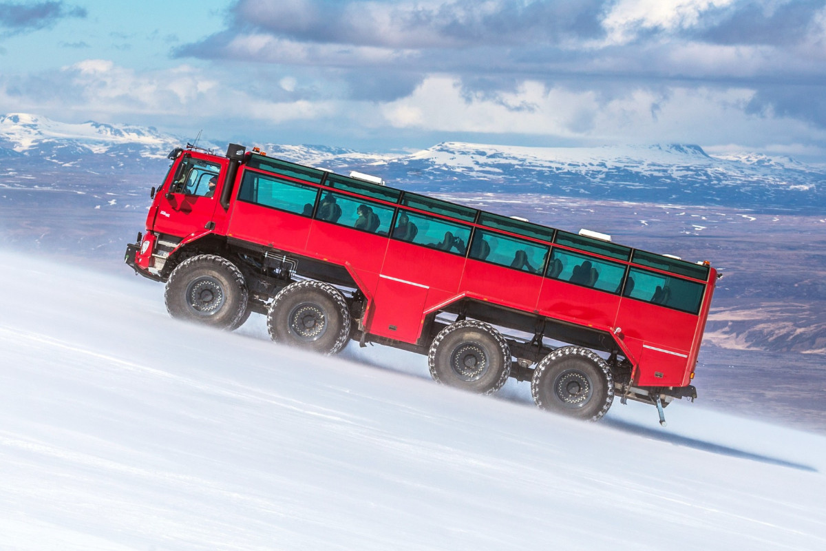 The impressive red Sleipnir monster truck ascending a snowy slope with mountains and a cloudy sky in the background during a glacier tour in Iceland.