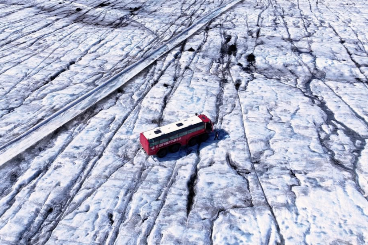 An aerial view of the large red Sleipnir monster truck parked on a vast, textured glacier in Iceland, showing the cracked ice patterns.