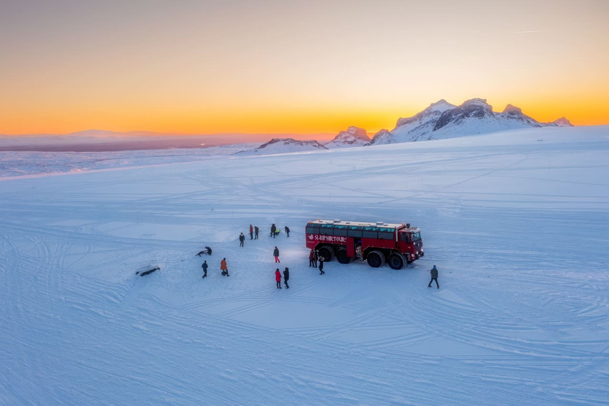 A group of people disembarking from the red Sleipnir monster truck on a vast snow-covered plain at sunset, with distant mountains illuminated by the orange sky in Iceland.